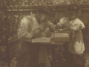 Fotografie welche Helene von Wild an einem niedrigen Tisch im Freien am Schreiben zeigt. Neben ihr steht ein Kleinkind, welches ein Papier vom auf dem Tisch liegenden Papierstapel hebt.