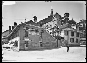Die Fotografie zeigt die Fassade des Restaurant Zähringer um 1913. Auf der Fassade sind diverse Werbeschilder angebracht auf welchen zu lesen ist: «Electrischer Aufzug Matte Plattform», «Achermann Tabak & Cigaren, Mercerie & Glaswaren», «Hess Bier».