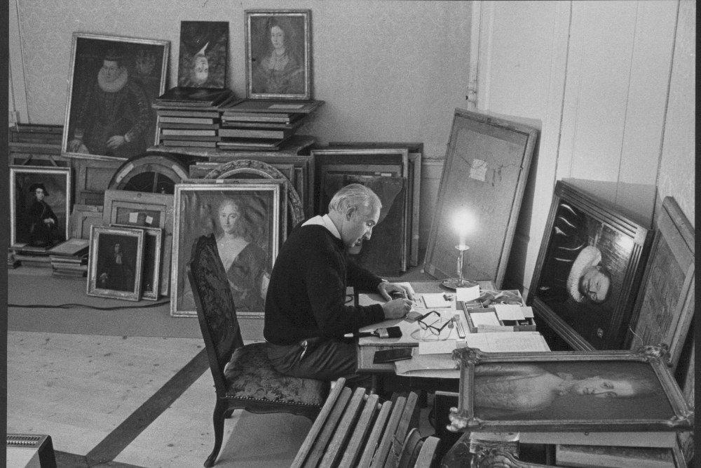 Fotografie zeigt einen Mann, welcher an einem Tisch arbeitet. Um in herum stapeln sich zahlreiche Gemälde.